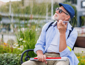 An older man on a bench talking on a phone with a white cane folded in his lap