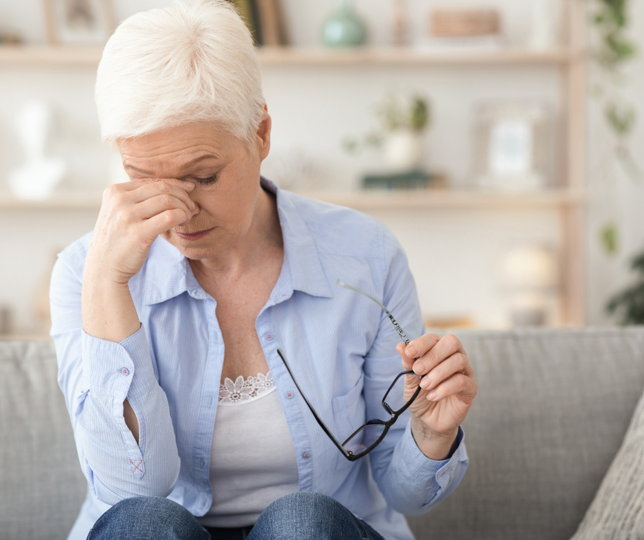 10 Signs of Age-Related Sight Loss - image of an older woman with short white hair, wearing a light blue cardigan, white blouse and blue jeans. She’s sitting with her head bowed and slightly resting on her right hand. Her eyes are closed and she’s holding her glasses with her left hand as if she’s taking a rest from using them. 