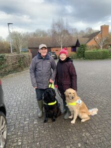 This article, Meet Margaret, image is a picture of Roger and Margaret. They are standing side by side in front of a lovely brick house. It’s an overcast day and they are wearing coats and hats. They’re each holding onto their guide dogs who are sitting in front of them.