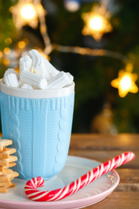 Sky blue cup on a white saucer filled with hot chocolate and topped with swirly whipped cream. Next to it is a candy cane and in the background is a Christmas tree and lights.