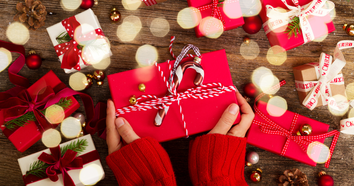 A gift wrapped in red wrapping paper and decorated with red and white, candy cane patterned ribbon, with a candy cane at the very center. The present is held, only the hands and part of the red knitted sweater sleeves are visible from the bottom of the image. The background has other finely wrapped and decorated gifts and Christmas lights. Article Image for “12 Stocking Stuffers and Gift Ideas for Blind and Visually Impaired People”