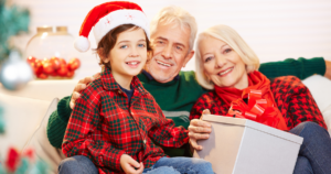 Grandparents sitting with a grandchild and holding a white package that’s about to be opened. They are all wearing holiday themed red and green outfits and the little boy is wearing a Santa hat - "Make Your Home Accessible for Holiday Visits"