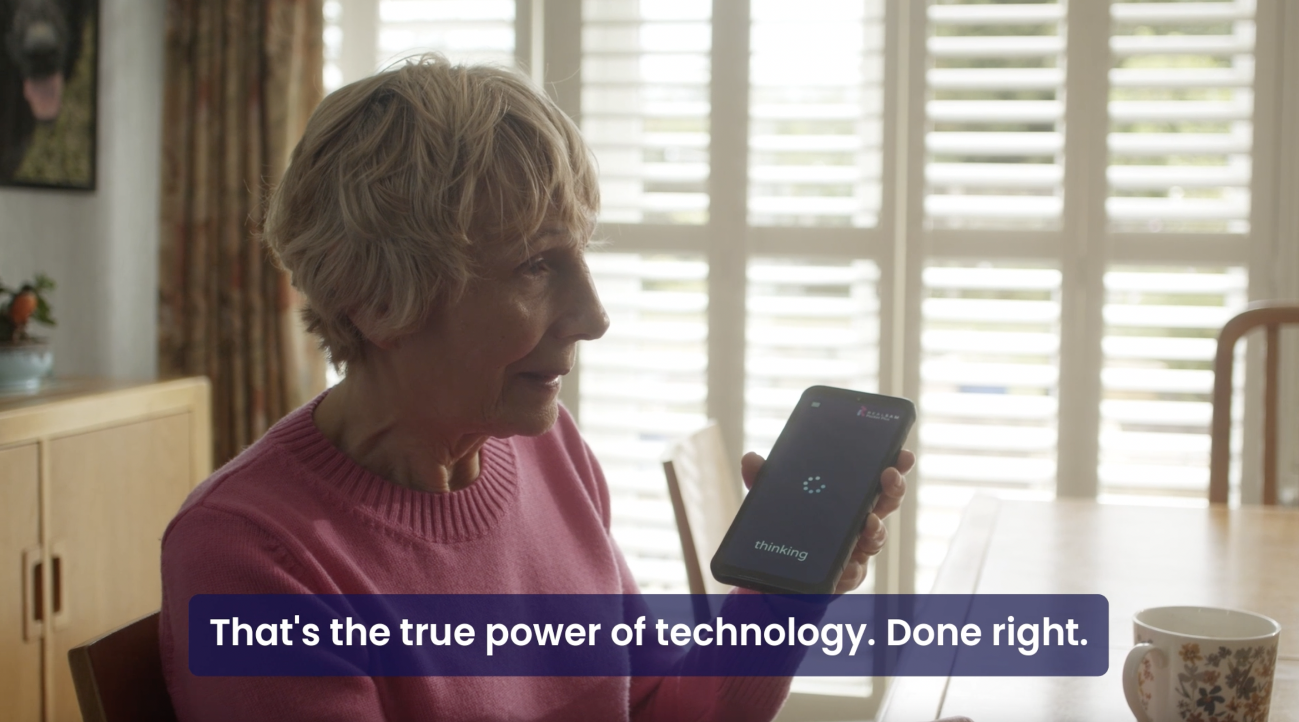 A smiling elderly woman in a pink sweater sits in a sunlit room holding a RealSAM device that displays the word "thinking." Text below reads, "That's the true power of technology. Done right,” identifying RealSAM as a powerful assistiev technology for vision loss.